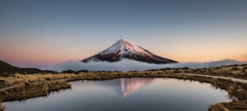 Sunrise over Mount Taranaki volcano with reflection in lake. Taranaki volcano with snow sorrounded by morning fog mirroring in water. Mirror pools at the Pouakai hut track in North island, New Zealand